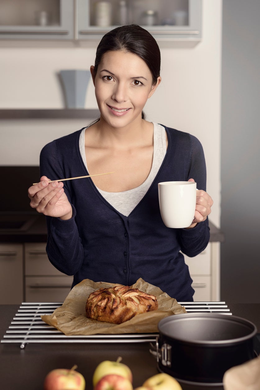 Smiling happy housewife baking in the kitchen standing over a freshly baked apple cake cooling on a rack with a mug of coffee in her hand